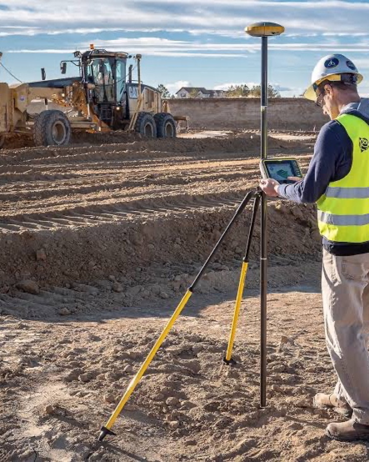 Trimble field technician using a GNSS rover beside a Caterpillar motor grader