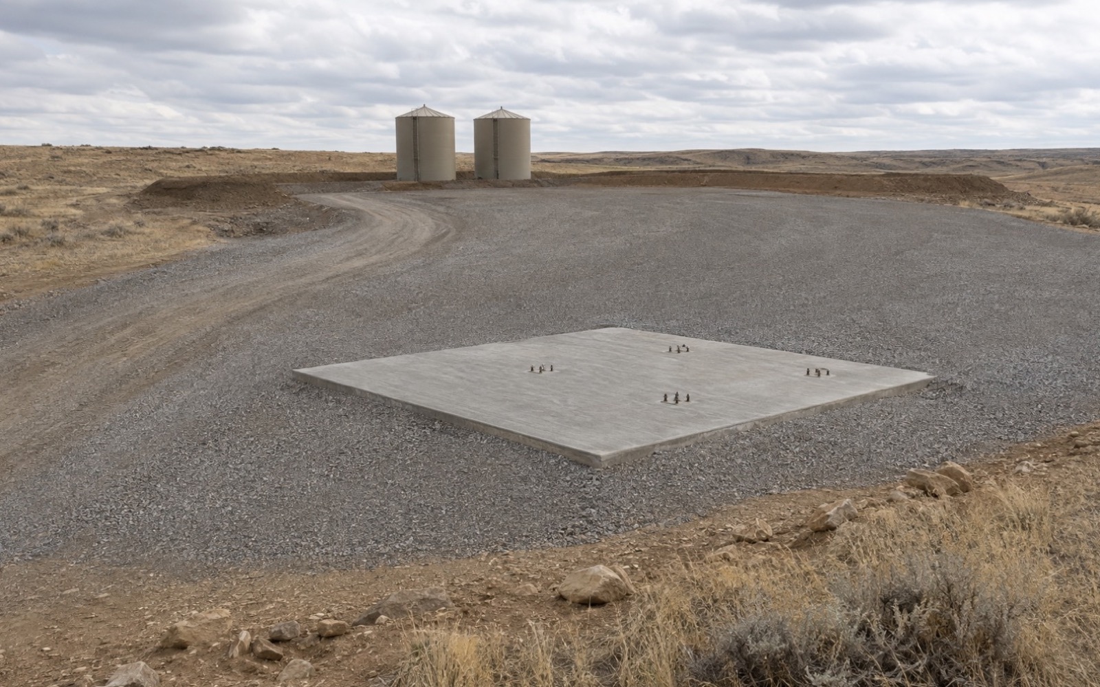 Illustrative oilfield construction scene with a gravel equipment pad, concrete foundation, access road, and distant storage tanks